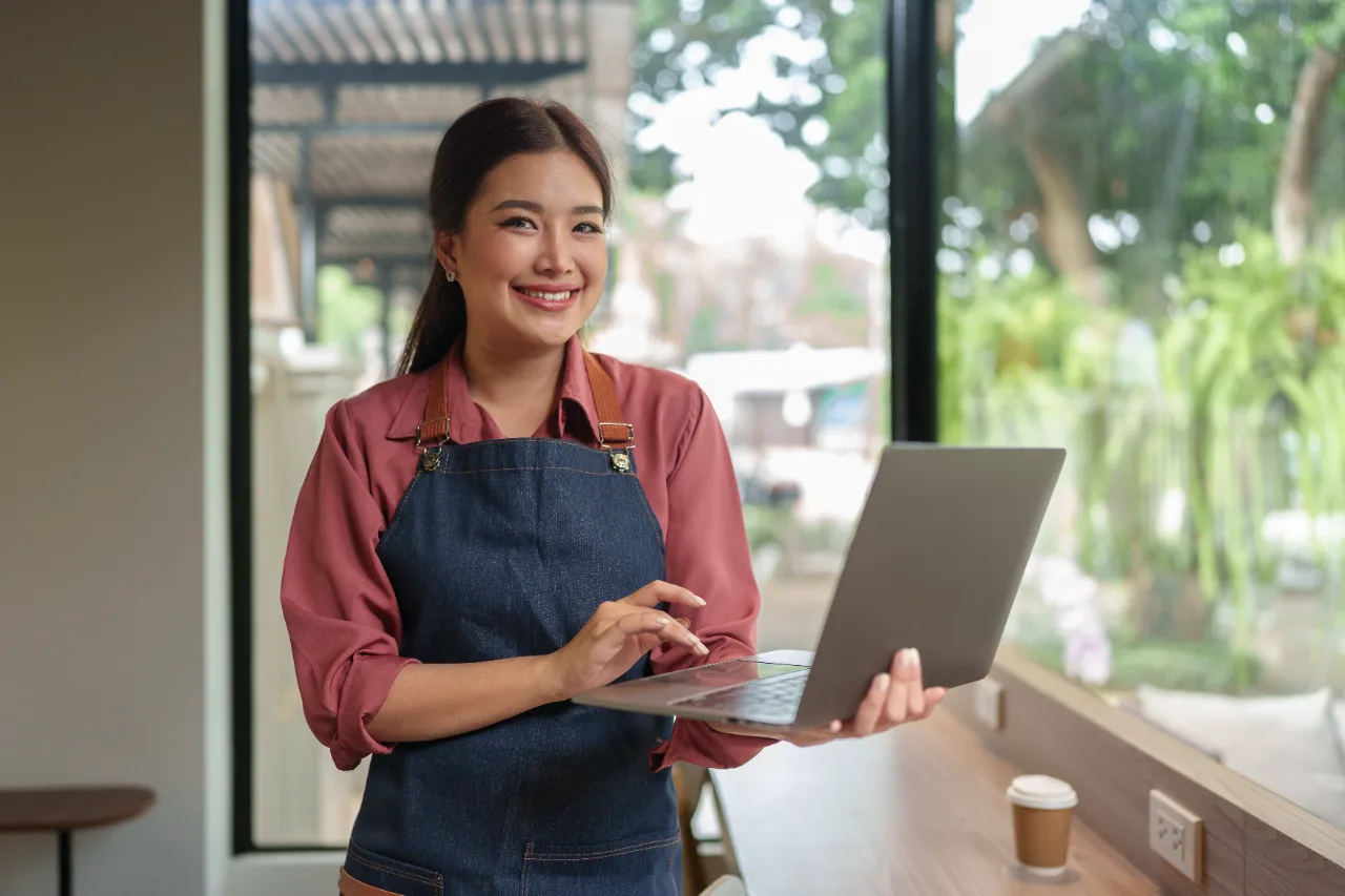 Smiling small-business owner in an apron working on a laptop in her café